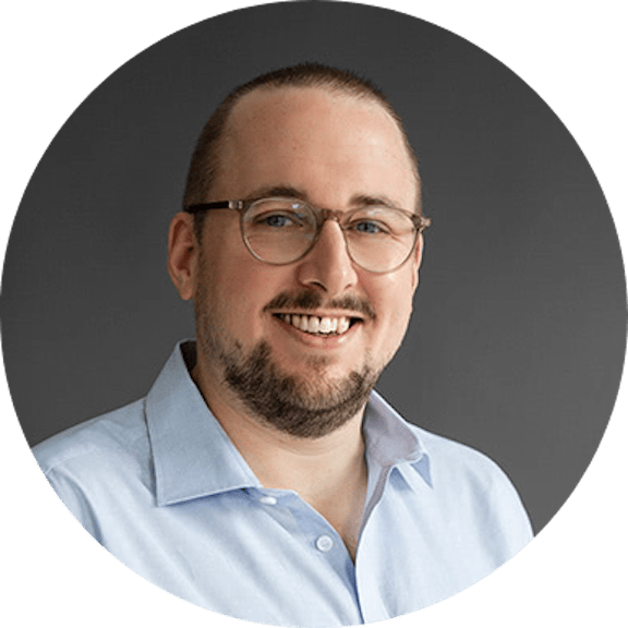 Porträt eines lächelnden Mannes mit Brille und hellblauem Hemd vor grauem Hintergrund