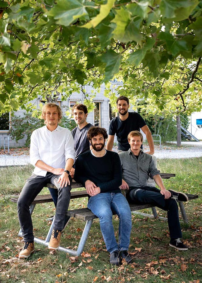 Five men sitting together on a bench under green foliage in front of a light-colored building, smiling at the camera