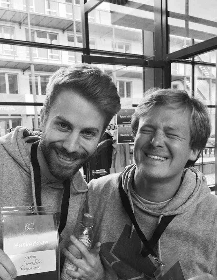 Hackerkiste Speaker Event Zwei Männer lächeln in die Kamera, einer trägt ein Hackerkiste-Speaker-Abzeichen bei einer Veranstaltung