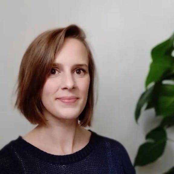 Portrait of a woman with brown shoulder-length hair smiling in front of white wall and green plant