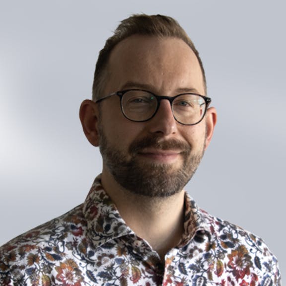 Portrait eines Mannes mit Brille und Vollbart, trägt ein gemustertes Hemd mit floralen Motiven