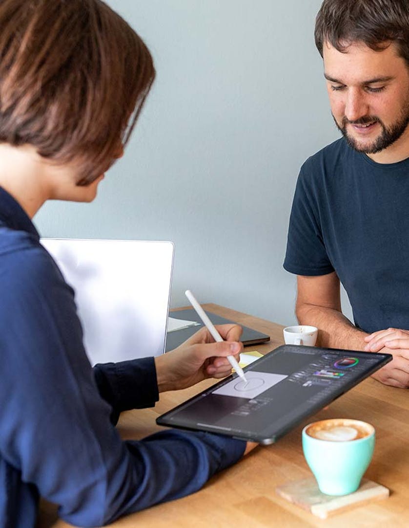 Two people sitting at a wooden table collaborating on a tablet displaying design software. One person holds a stylus while the other looks on, with laptops and coffee cups visible.