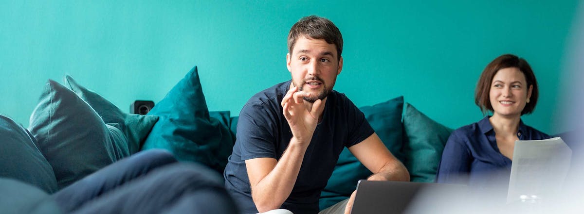 Two Peeris sitting on a couch with turquoise background, collaborating on laptops during the Weekly Meeting