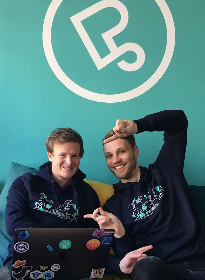 Two Peeris in dark hoodies with Peerigon logo sit in front of turquoise wall with Peerigon symbol, holding a sticker-covered laptop