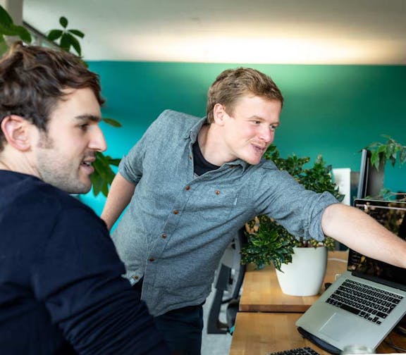 Teamarbeit im Peerigon-Büro Zwei Peerigon-Entwickler schauen lachend auf einen Laptop-Bildschirm in einem modernen Büro