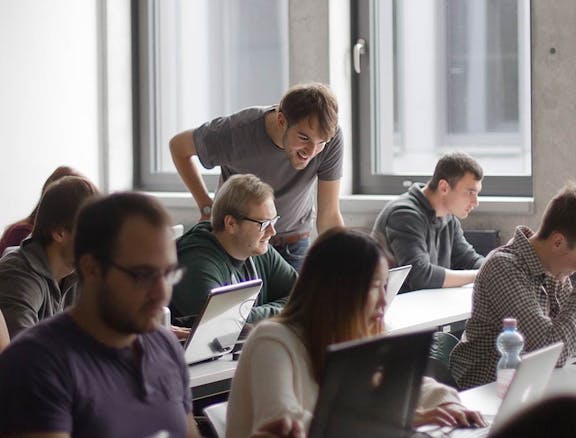 Teilnehmer in einem Workshop sitzen an Tischen mit Laptops, ein Peerigon-Trainer beugt sich über einen Kollegen, um ihm zu helfen.
