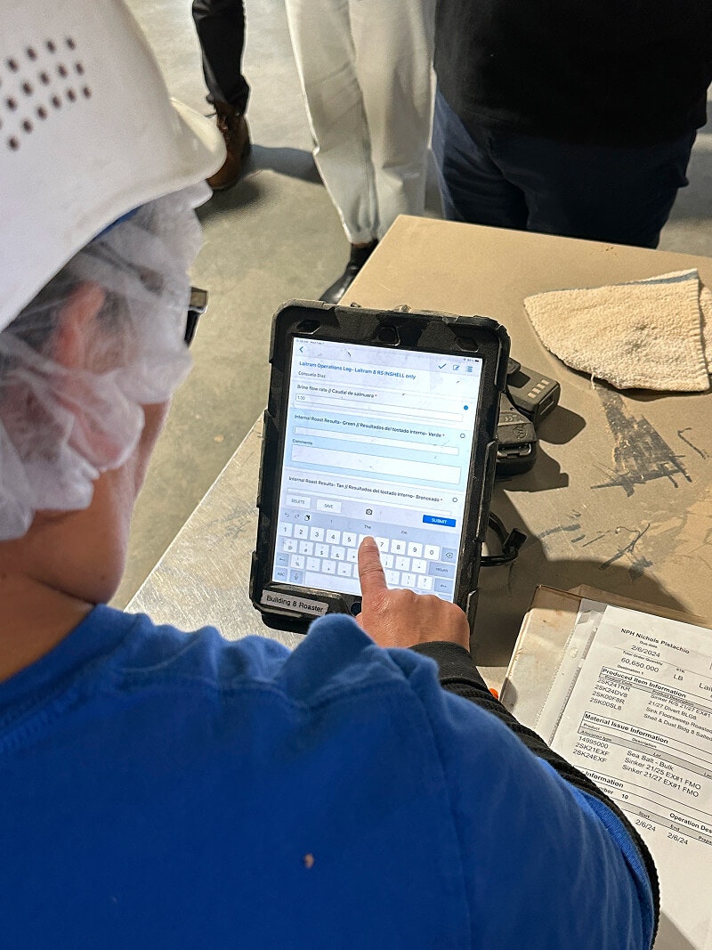 Worker in a food manufacturing facility using a tablet to input or review operational data, with visible safety gear and process documents on the table.