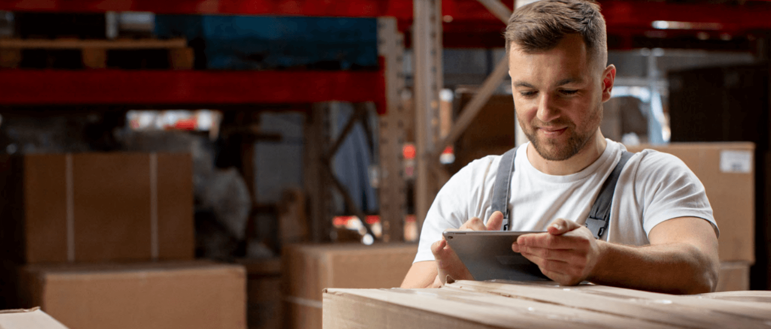 A man wearing overalls is standing in a warehouse, smiling while using a tablet. Large cardboard boxes are stacked around him, and shelves with more boxes are visible in the background.