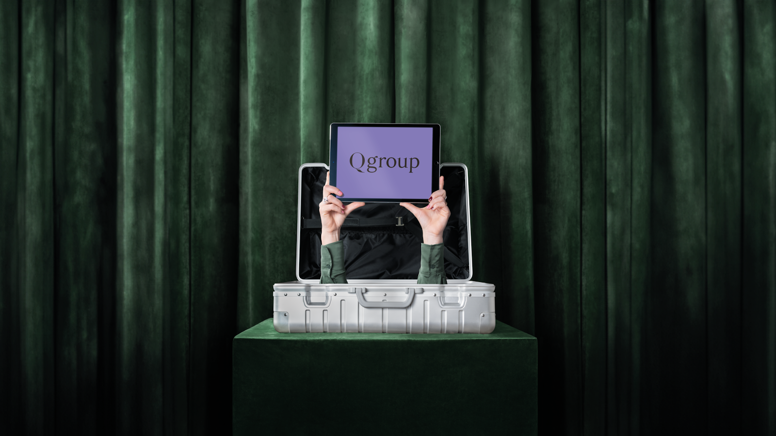 Hands emerging from a silver suitcase holding an iPad displaying the text 'Qgroup' on a purple background.
