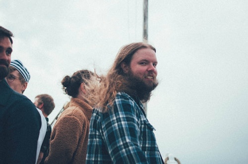 Man with long hair and plaid shirt smiling, surrounded by people on a boat.