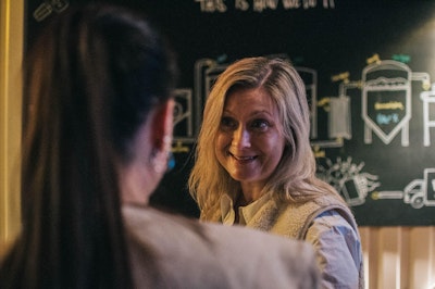 A woman with long blonde hair smiles while talking to another person in a dimly lit setting.