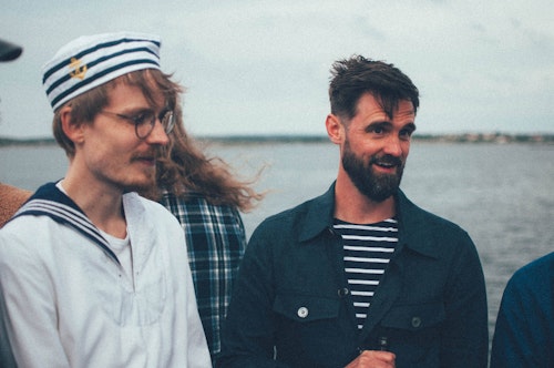 Two men by the water; one in a sailor outfit, the other in a striped shirt.
