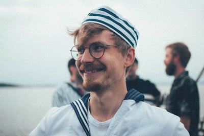Smiling man in a sailor hat and glasses, with a blurred background of people and water.