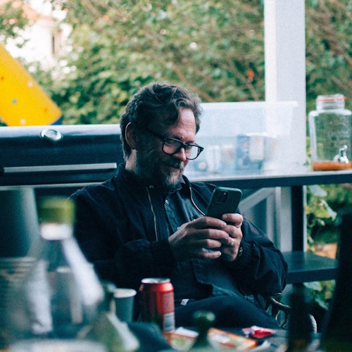 Man with glasses sitting outdoors, looking at his phone, surrounded by drinks and greenery.