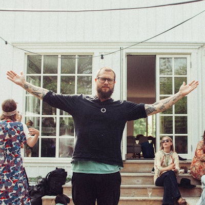 Man with tattoos and glasses stands with arms outstretched in front of a house, people socializing in the background.