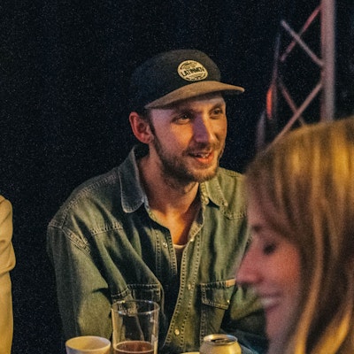 Man in a cap smiling while talking to a woman with long hair, both in a dimly lit setting.