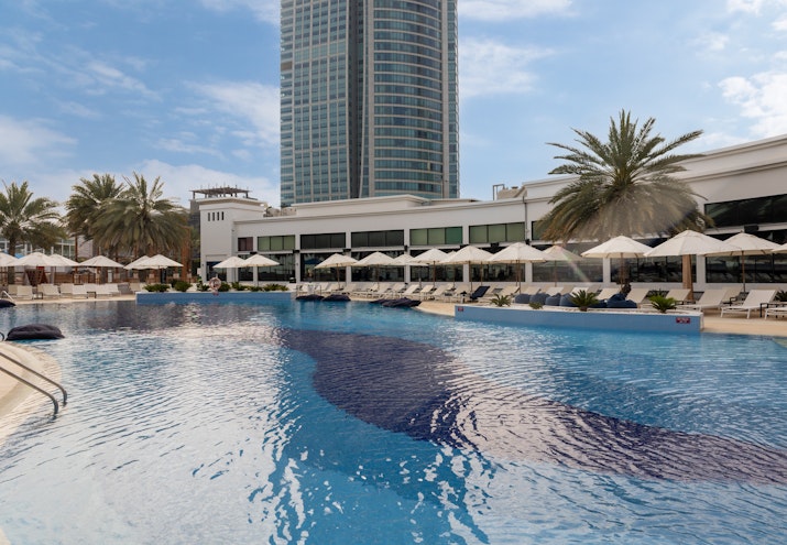 West Bay Abu Dhabi A pool with sunbeds and umbrellas, with a modern building and palm trees surrounding it, under a partly cloudy sky.