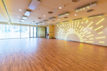 Empty fitness studio featuring wooden floors, large windows with city views, and a wall with sunburst LED lighting.