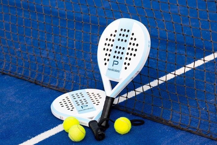 Two padel rackets resting against the net on a vibrant blue padel court with three yellow balls scattered nearby, ready for play