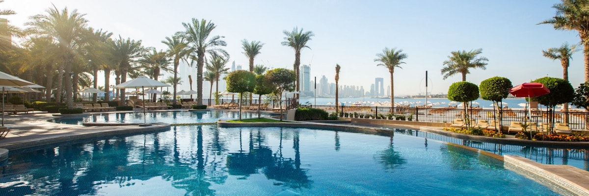 Fairmont The Palm A clear blue pool surrounded by palm trees and lounge chairs, with a view of a city skyline in the background.