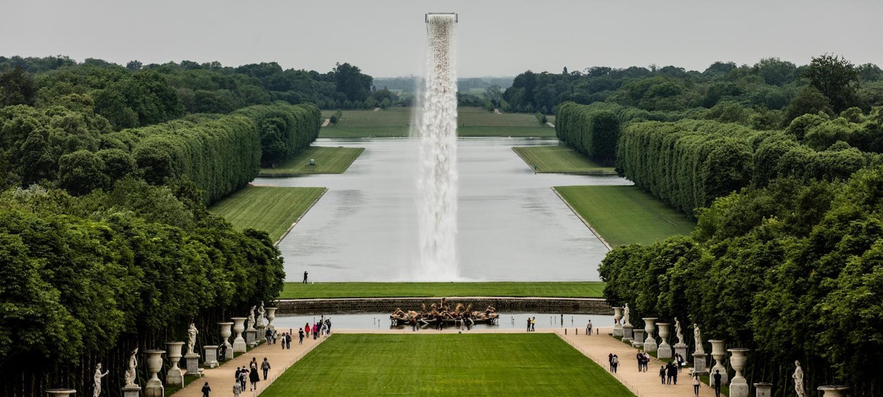 Eine weite Ansicht der formalen Gärten von Versailles mit einem langen spiegelnden Teich, gepflegten Rasenflächen, ordentlich geschnittenen Hecken, einem zentralen Brunnen mit einem hohen Wasserstrahl und Besuchern, die die Wege entlanggehen.
