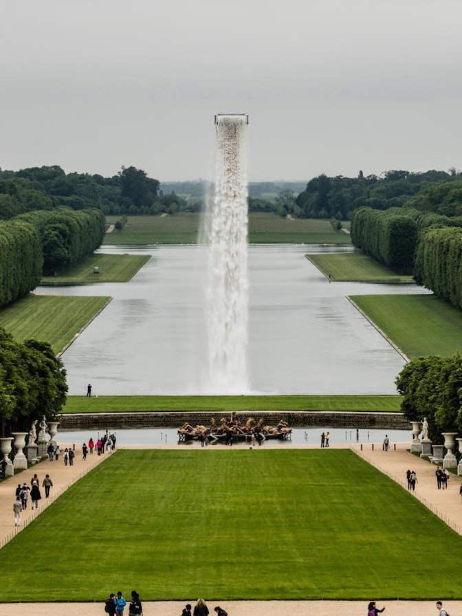 Waterfall Olafur Eliasson Eine weite Ansicht der formalen Gärten von Versailles mit einem langen spiegelnden Teich, gepflegten Rasenflächen, ordentlich geschnittenen Hecken, einem zentralen Brunnen mit einem hohen Wasserstrahl und Besuchern, die die Wege entlanggehen.