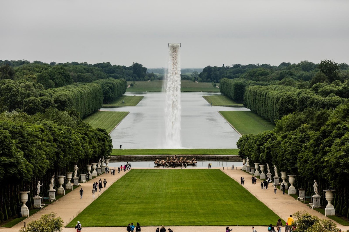 Eine weite Ansicht der formalen Gärten von Versailles mit einem langen spiegelnden Teich, gepflegten Rasenflächen, ordentlich geschnittenen Hecken, einem zentralen Brunnen mit einem hohen Wasserstrahl und Besuchern, die die Wege entlanggehen.