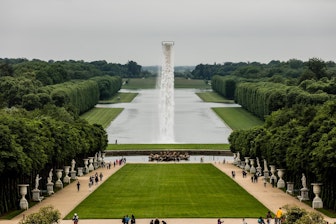 Eine weite Ansicht der formalen Gärten von Versailles mit einem langen spiegelnden Teich, gepflegten Rasenflächen, ordentlich geschnittenen Hecken, einem zentralen Brunnen mit einem hohen Wasserstrahl und Besuchern, die die Wege entlanggehen.