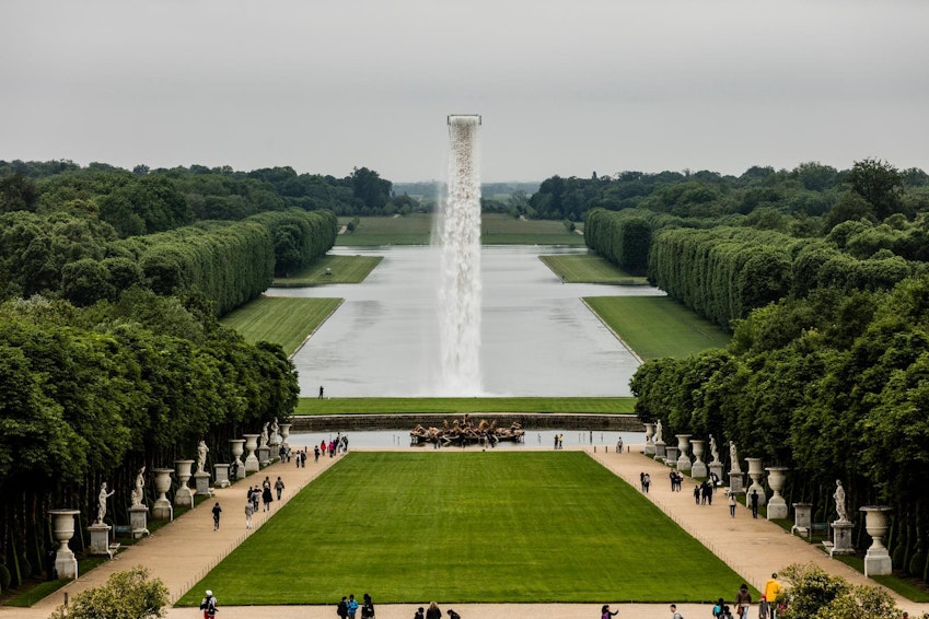 Eine weite Ansicht der formalen Gärten von Versailles mit einem langen spiegelnden Teich, gepflegten Rasenflächen, ordentlich geschnittenen Hecken, einem zentralen Brunnen mit einem hohen Wasserstrahl und Besuchern, die die Wege entlanggehen.
