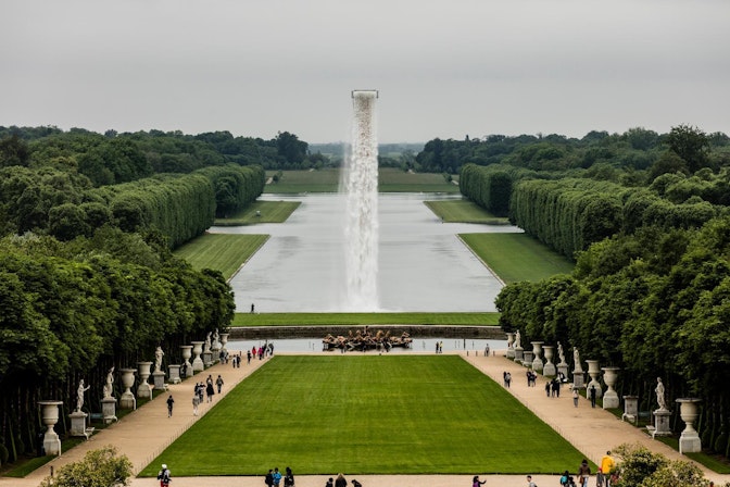Eine weite Ansicht der formalen Gärten von Versailles mit einem langen spiegelnden Teich, gepflegten Rasenflächen, ordentlich geschnittenen Hecken, einem zentralen Brunnen mit einem hohen Wasserstrahl und Besuchern, die die Wege entlanggehen.
