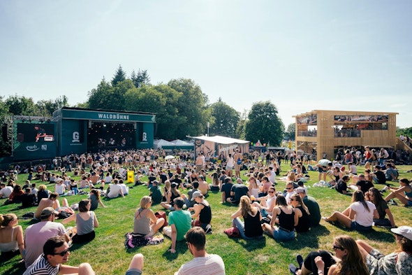Pubblico sul prato davanti alla Waldbühne al Gurtenfestival, con il padiglione in legno a due piani sulla destra