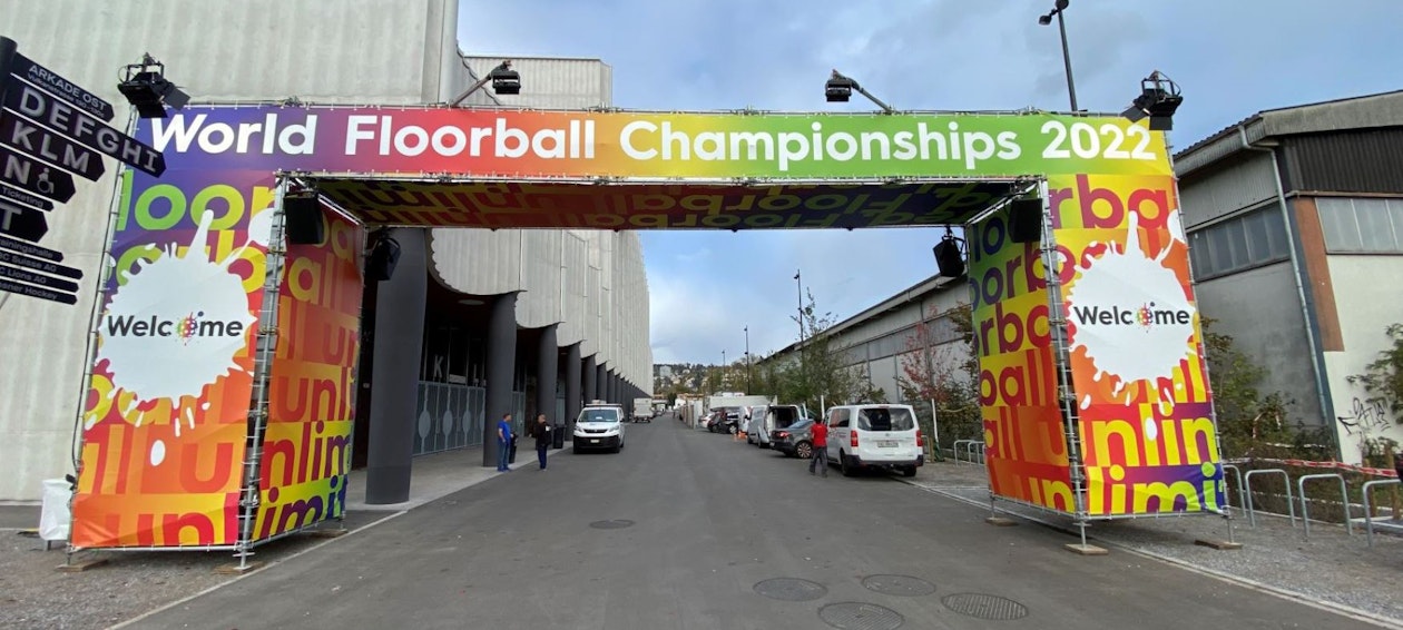World Floorball Championships Ein farbenfroher Torbogen mit den Schildern Unihockey-WM 2022 und Willkommen steht über einer Straße, die zu einem Veranstaltungsort mit Menschen und geparkten Fahrzeugen in der Nähe führt.