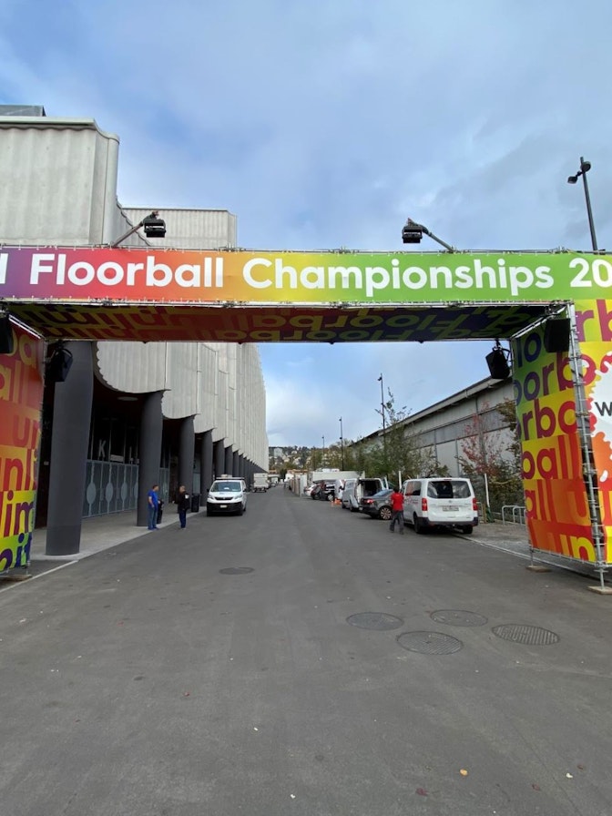 Unihockey Weltmeisterschaft Ein farbenfroher Torbogen mit den Schildern Unihockey-WM 2022 und Willkommen steht über einer Straße, die zu einem Veranstaltungsort mit Menschen und geparkten Fahrzeugen in der Nähe führt.