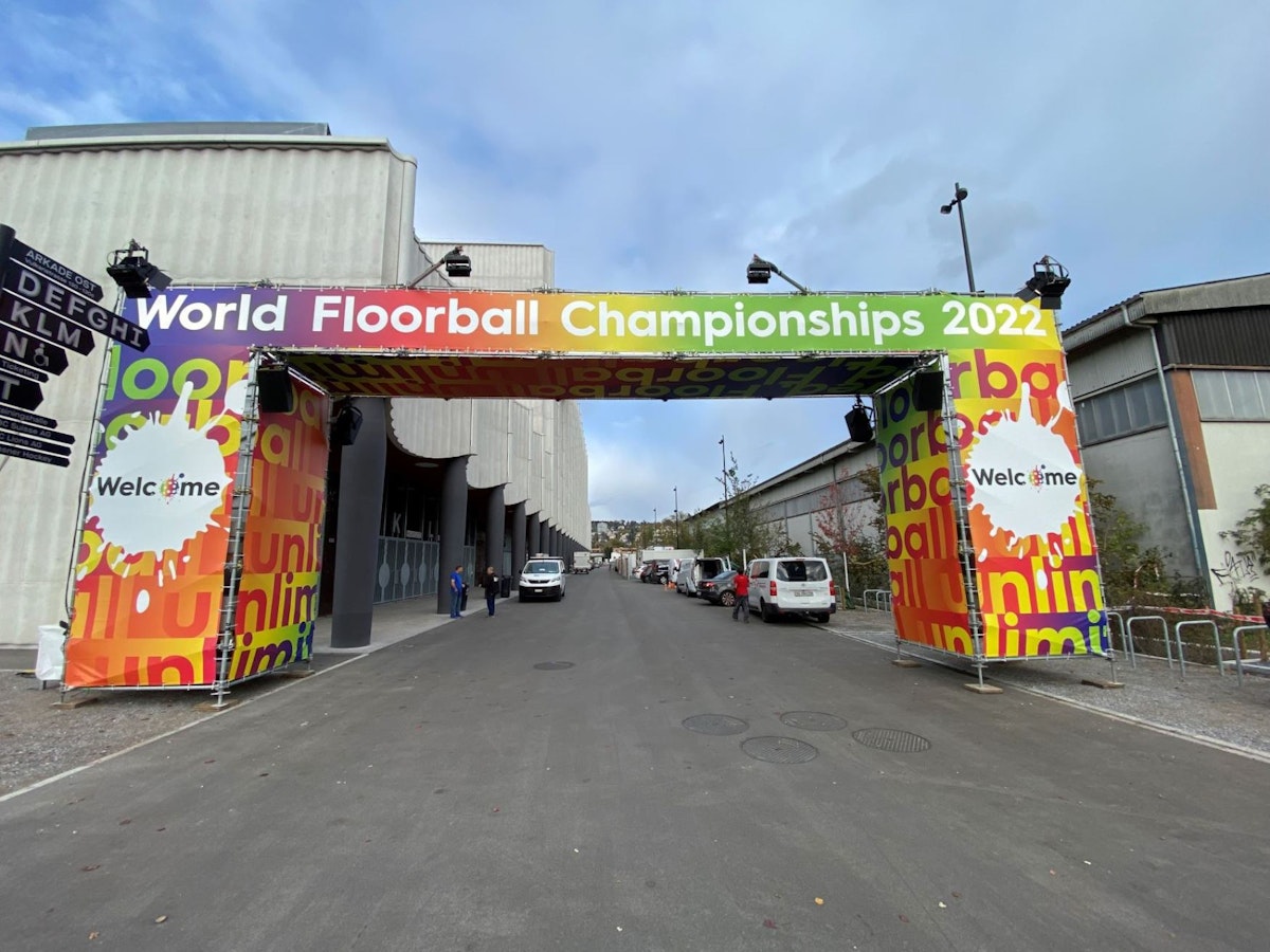 Ein farbenfroher Torbogen mit den Schildern Unihockey-WM 2022 und Willkommen steht über einer Straße, die zu einem Veranstaltungsort mit Menschen und geparkten Fahrzeugen in der Nähe führt.