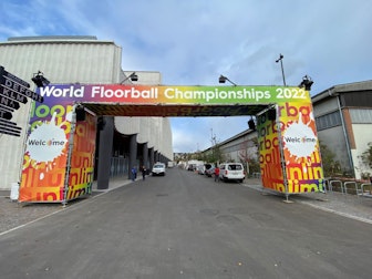 Ein farbenfroher Torbogen mit den Schildern Unihockey-WM 2022 und Willkommen steht über einer Straße, die zu einem Veranstaltungsort mit Menschen und geparkten Fahrzeugen in der Nähe führt.