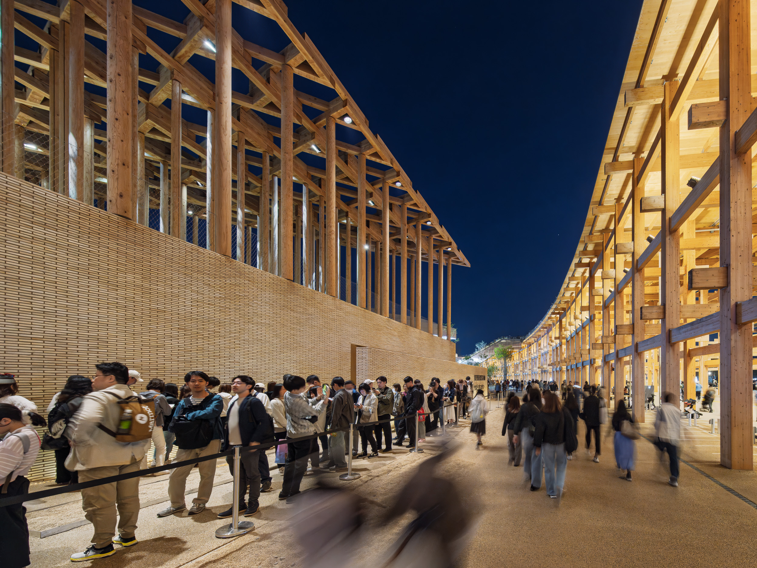 Los visitantes esperan por la noche frente al pabellón de madera iluminado de la Expo 2025 en Japón.