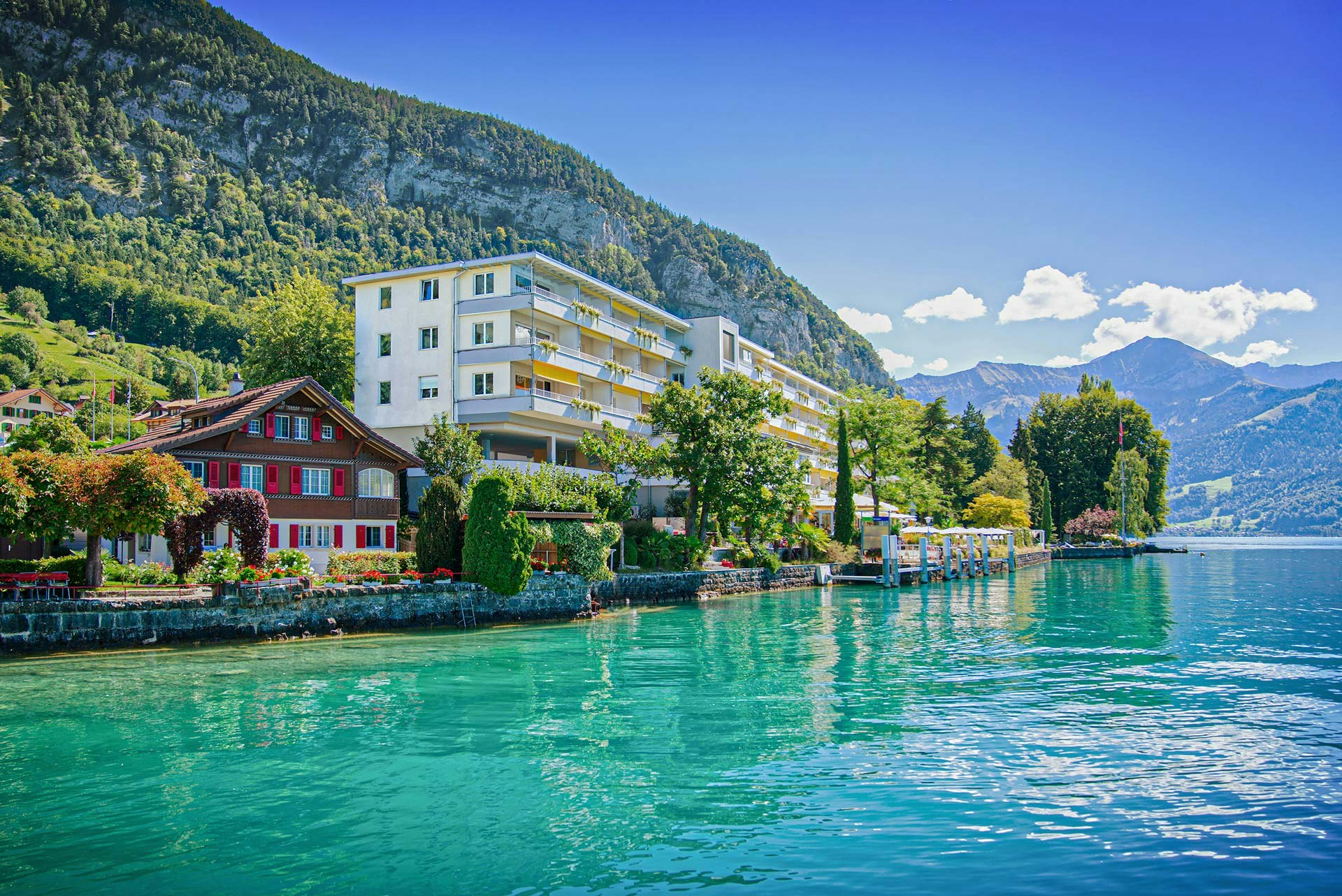 Sonniger Tag, türkisfarbener See, Hotelgebäude mit gelben Sonnenstoren vor Bergen