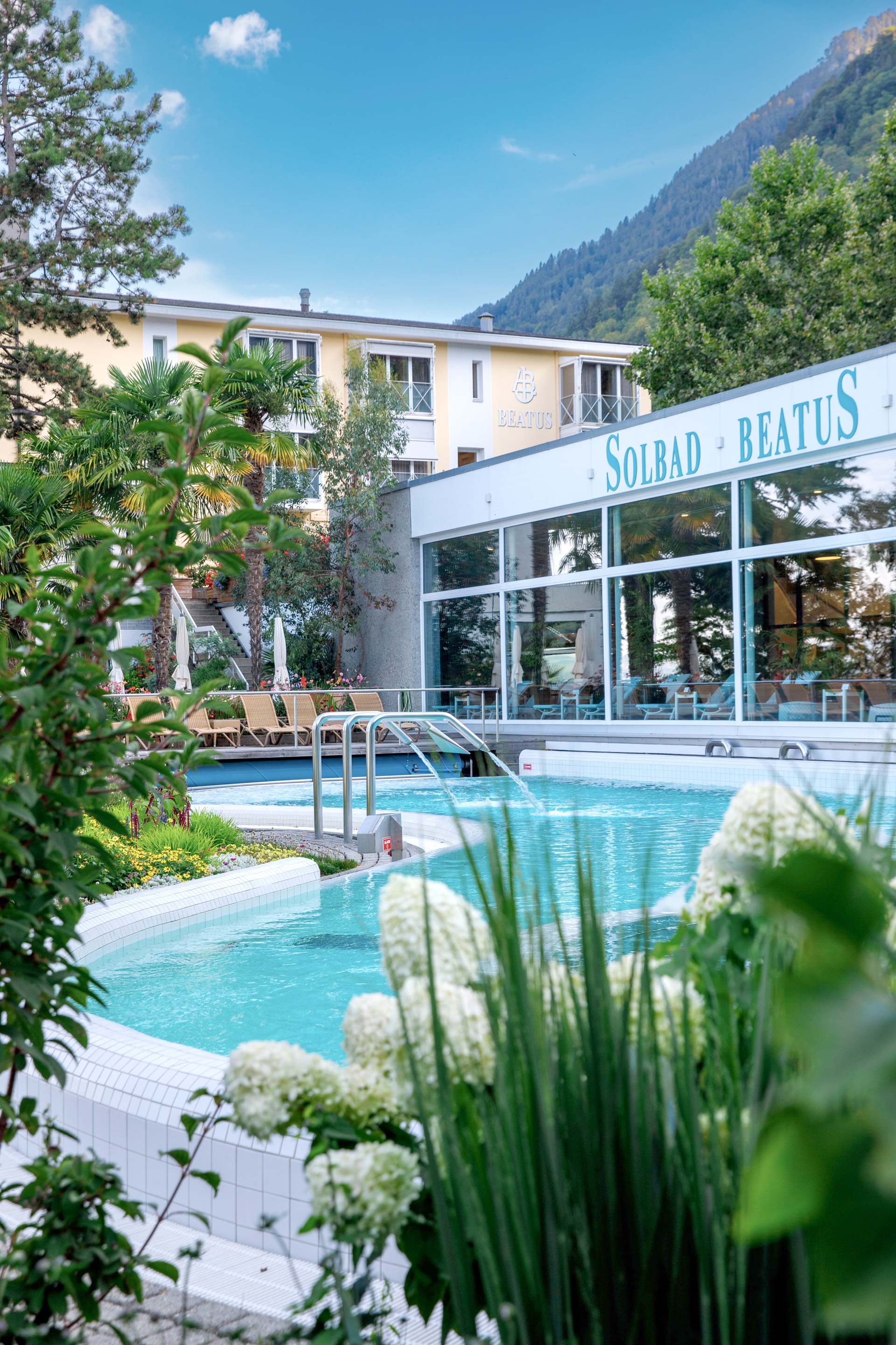 Blumen und Parklandschaft, links das Hotel Beatus, rechts die Schwimmhalle und das Aussenbecken