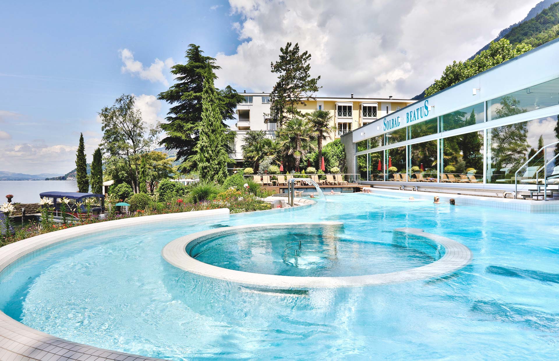 Im Vordergrund ein Schwimmbad mit hellblauem Wasser, im Hintergrund Bäume und das Hotel Beatus