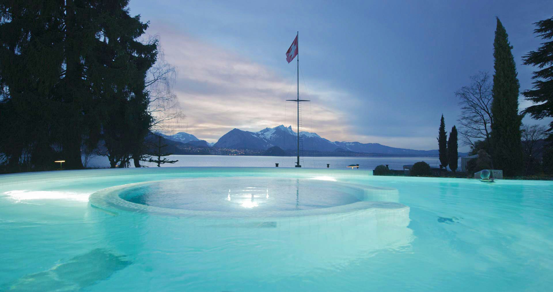 Beleuchtetes Schwimmbad, Schweizerfahne, ein kleiner Streifen des Thunersees und Berge in der Dämmerung
