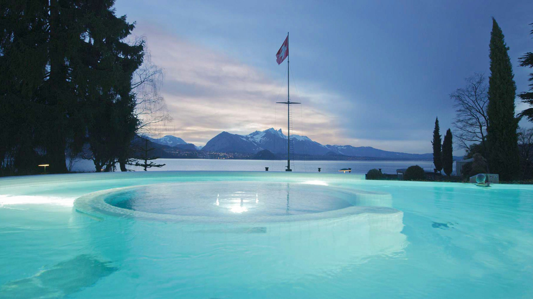 Solbad des Hotel BEATUS in der Dämmerung Beleuchtetes Schwimmbad, Schweizerfahne, ein kleiner Streifen des Thunersees und Berge in der Dämmerung