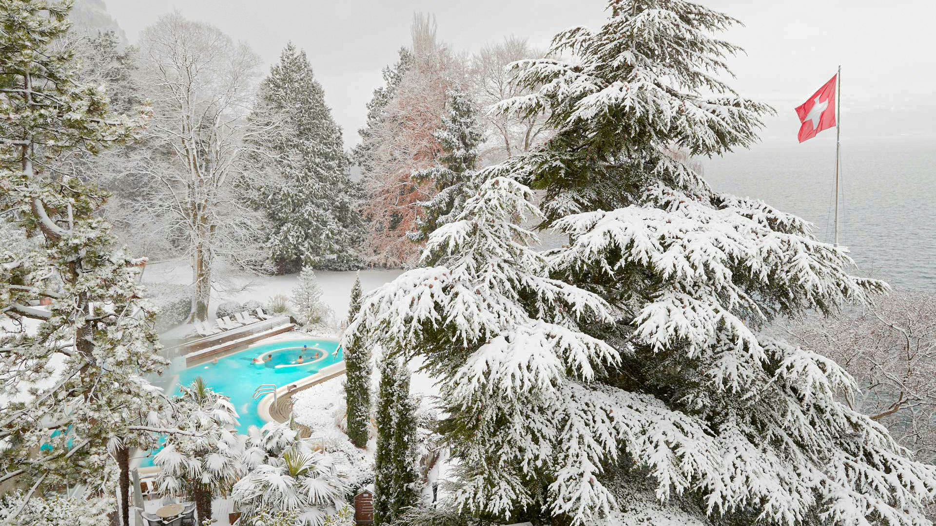 Solbad im Winter Verschneite Landschaft von schräg oben, links im Bild ein türkisfarbenes Schwimmbad