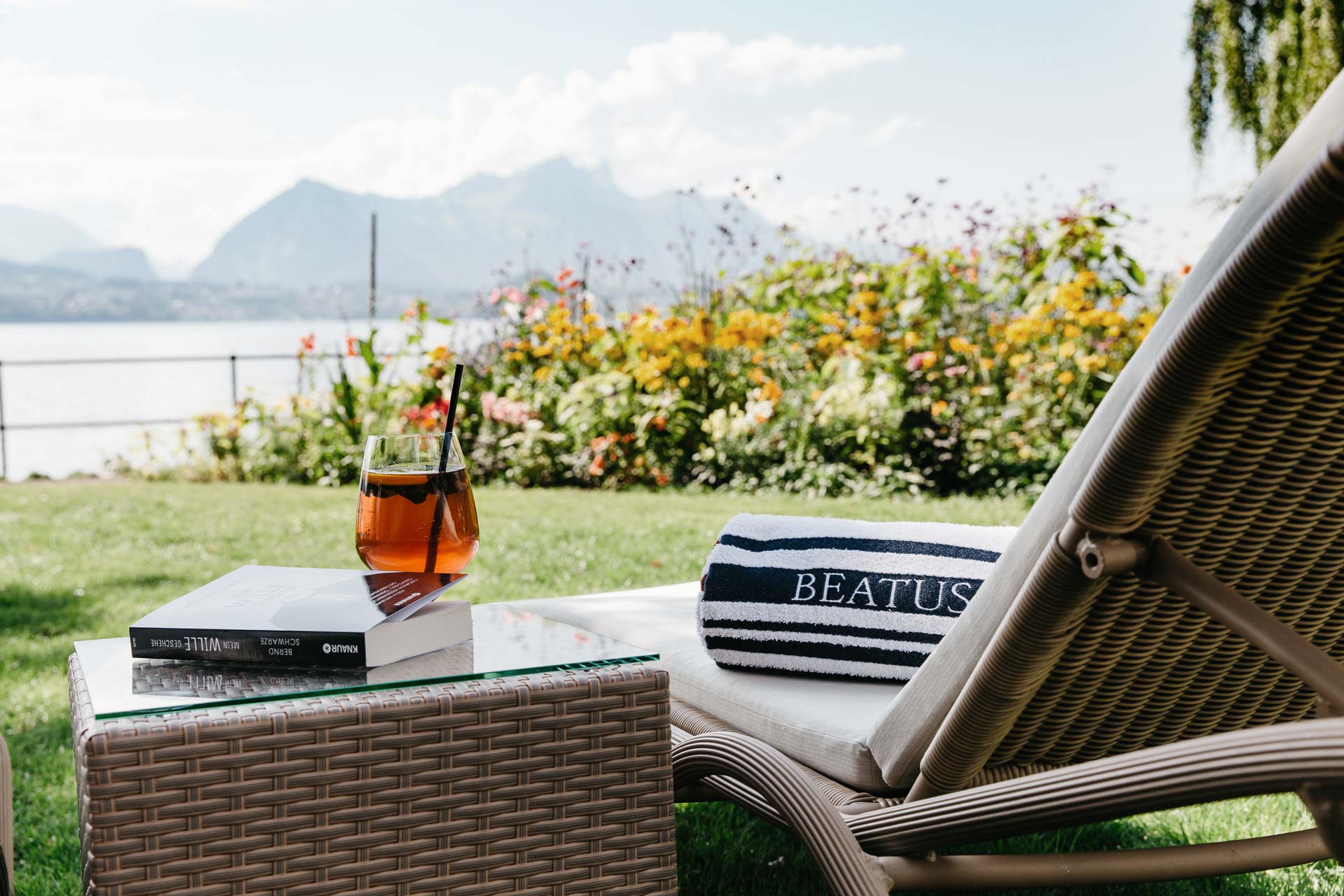 Sun lounger with bath towel, next to it a wicker table with aperitif and book, behind it the garden, lake and mountains
