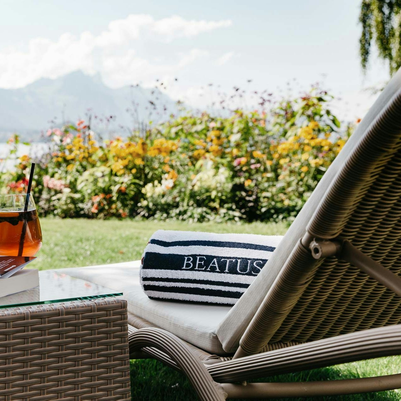 Chaise longue et apéritif dans le jardin de l'hôtel BEATUS Chaise longue avec serviette de bain, à côté d'une table en osier avec apéro et livre, derrière le jardin, le lac et les montagnes