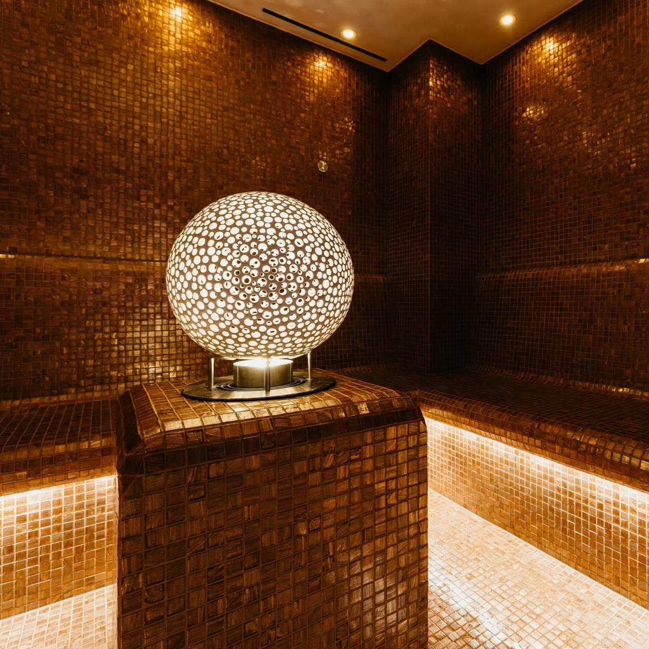 Light ball in the Aeris steam bath at Hotel BEATUS Light sphere with small brown and white dots on a plinth, room covered with brown mosaic tiles