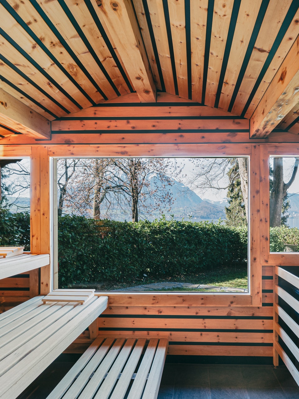 Schöne Sicht auf den Hotelpark von der Aussensauna des Hotel BEATUS Drei Niveaus von Holzliegen, Holzdecke, rechts ein Ofen mit Kohlen, grosses Panoramafenster mit Sicht auf den Hotelpark
