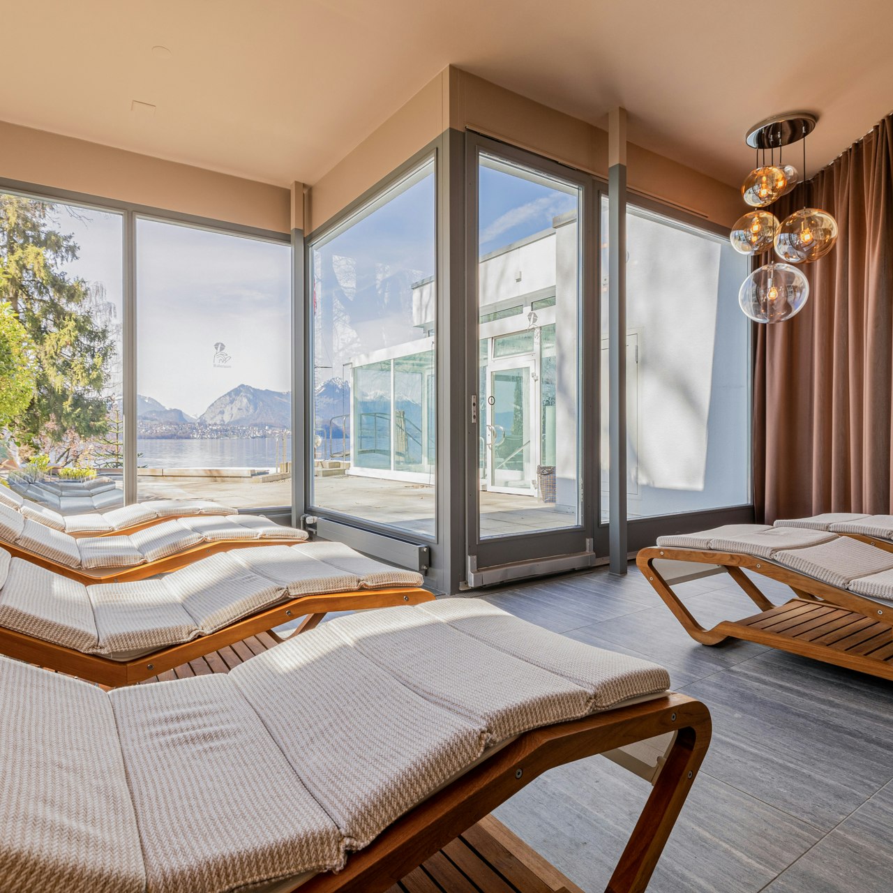 Salle de repos dans le sauna classique de l'hôtel BEATUS Chaises longues moelleusement rembourrées, derrière une terrasse ensoleillée donnant sur le parc et le lac