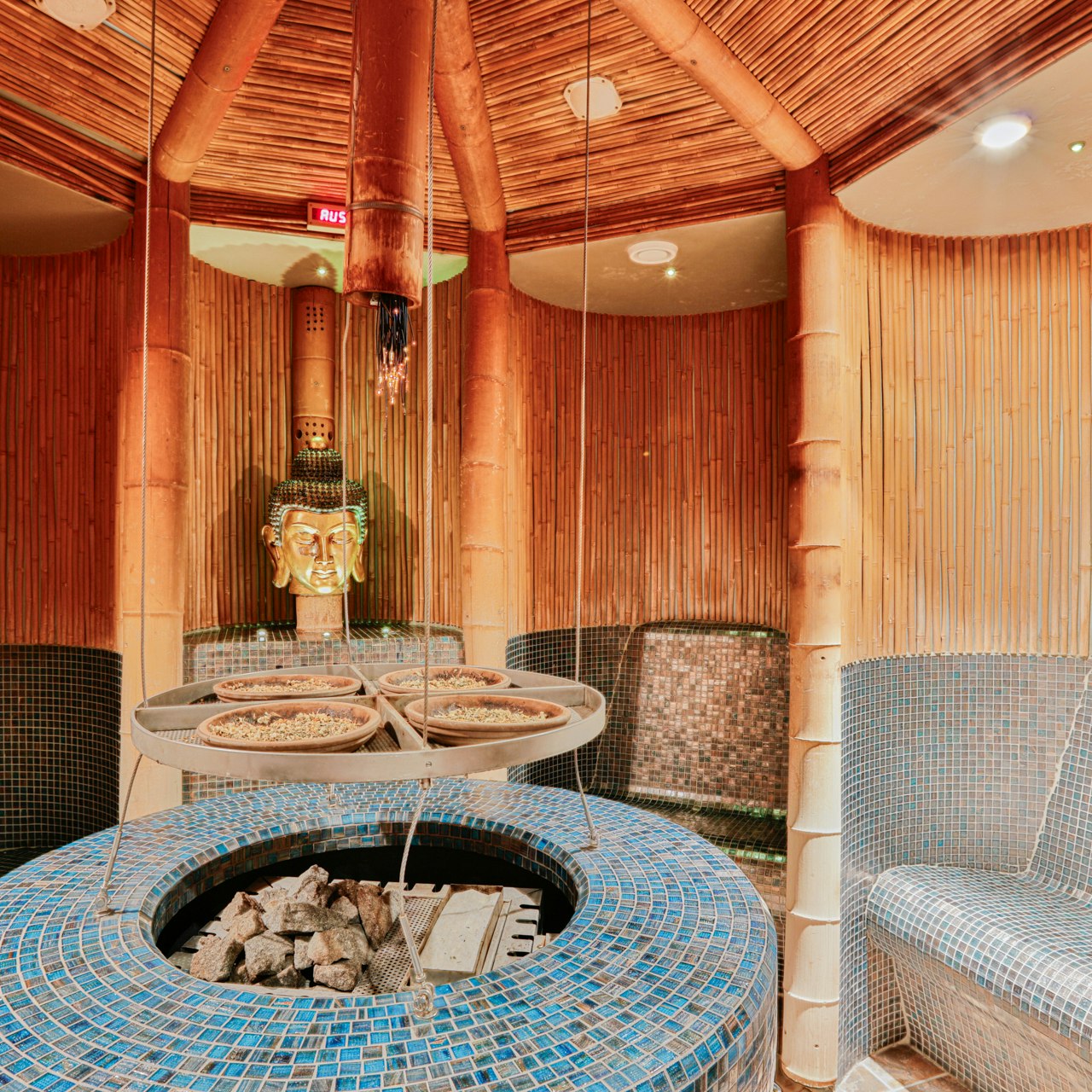 Sauna thaïlandais aux herbes à l'hôtel BEATUS Sauna avec poêle à infusion au centre, à gauche et à droite cabines à sièges revêtues de mosaïques en brun ou en bleu, à l'arrière une figure de Bouddha