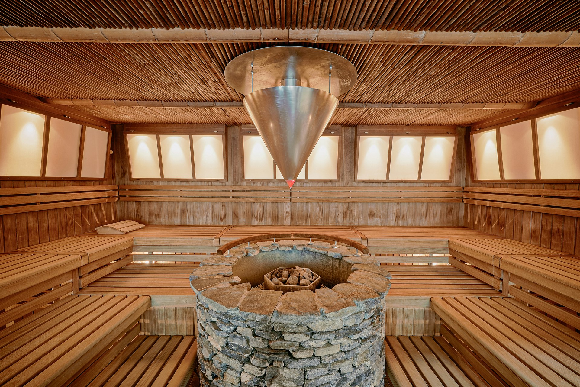 Large metal cone for automatic infusion over a round stone sauna heater, benches all round, small panoramic windows