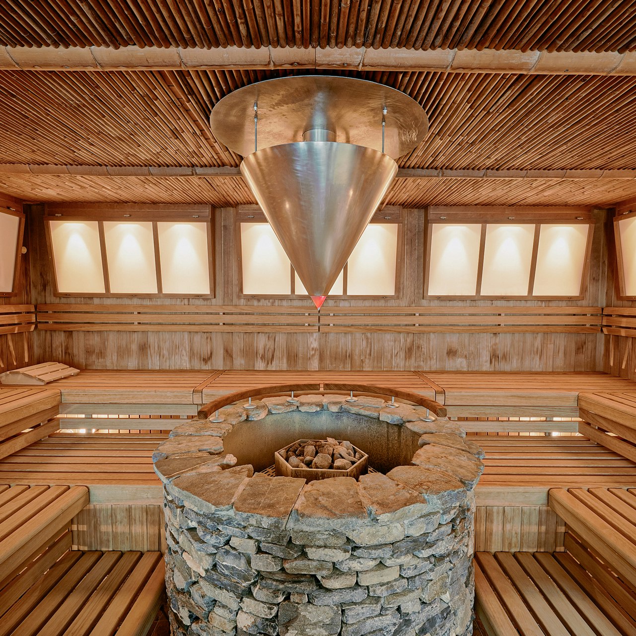 Sauna zen à l'hôtel BEATUS Grand cône métallique pour l'arrosage automatique au-dessus d'un poêle circulaire en pierre, bancs tout autour, petites fenêtres panoramiques